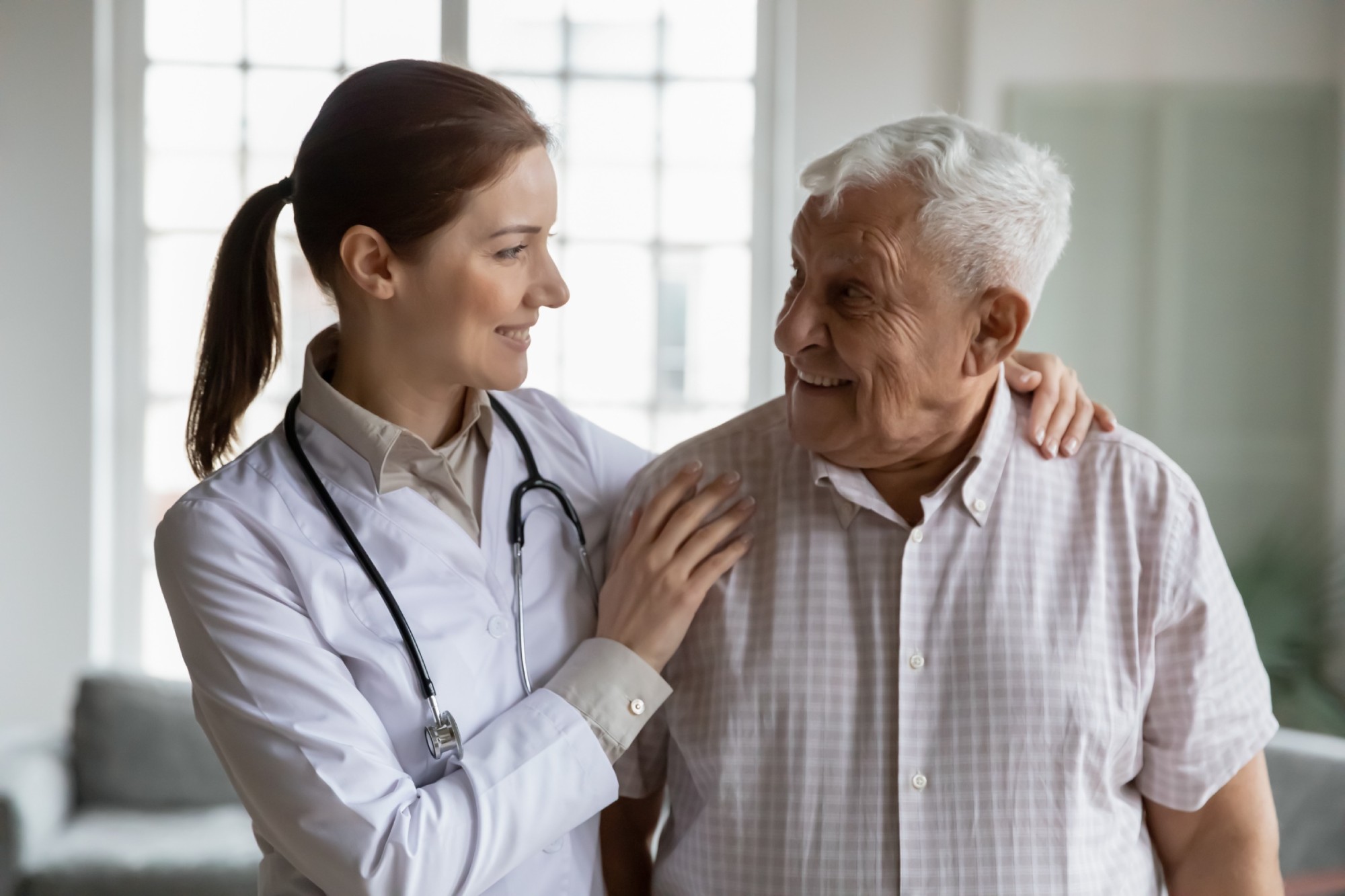 Head shot portrait smiling caring female doctor hugging older man, standing together, young woman caregiver wearing white coat and stethoscope supporting mature senior patient